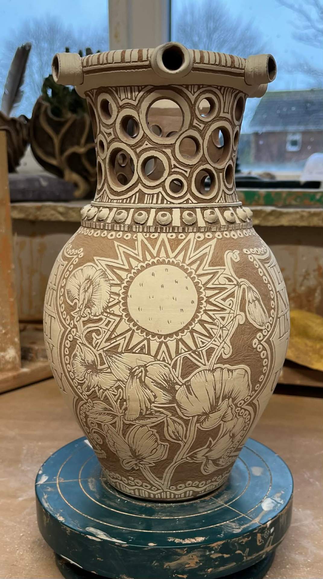 An unglazed carved sgraffito pot standing on the banding wheel in the studio, the whole pot in view.